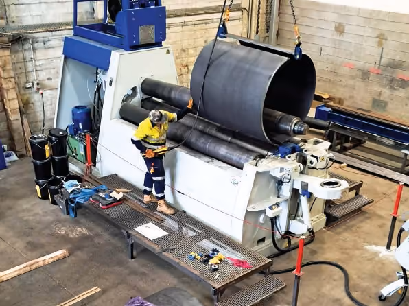 An ISS worker operates the plate roller by hand control while a finished steel cylinder hangs from a crane hook alongside.