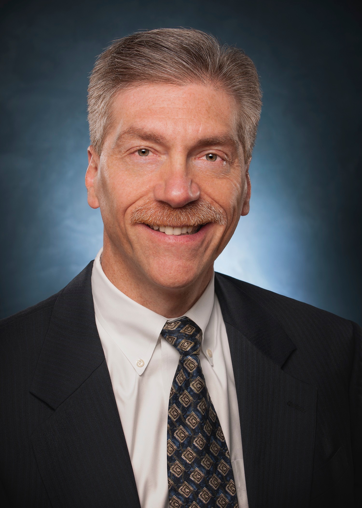Professional headshot of a Ken Senser in a dark suit and patterned tie, wearing a white shirt. He has short gray hair, a mustache, and is smiling against a dark gradient background.