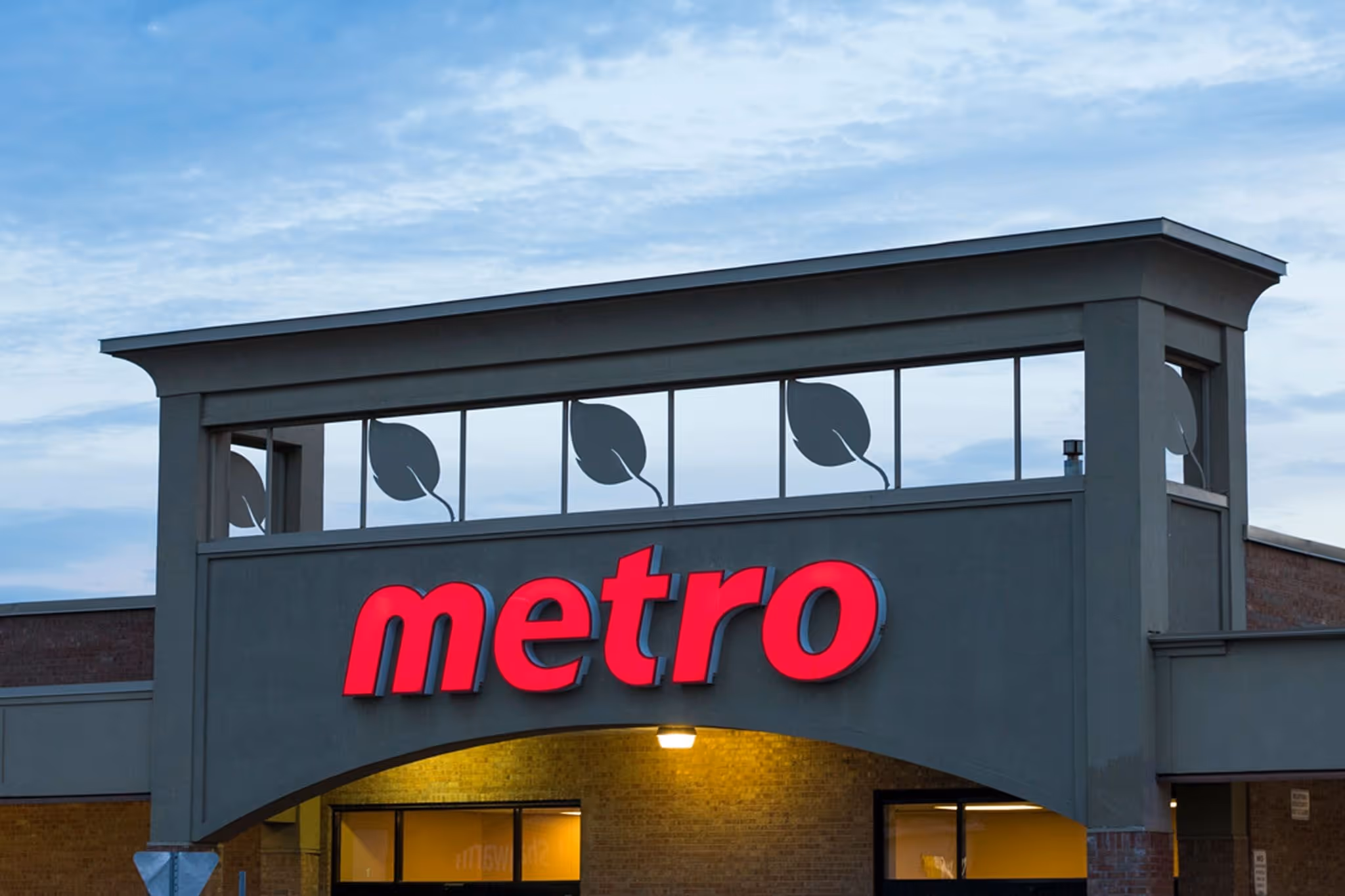 The image shows the exterior of a Metro grocery store, bustling with customers entering and exiting, eager to explore a variety of fresh fruits, vegetables, and deli prepared meals. The store's signage highlights its focus on quality organic groceries and everyday deals, inviting shoppers to enjoy a diverse selection of products, including meat, dairy, and health beauty items.