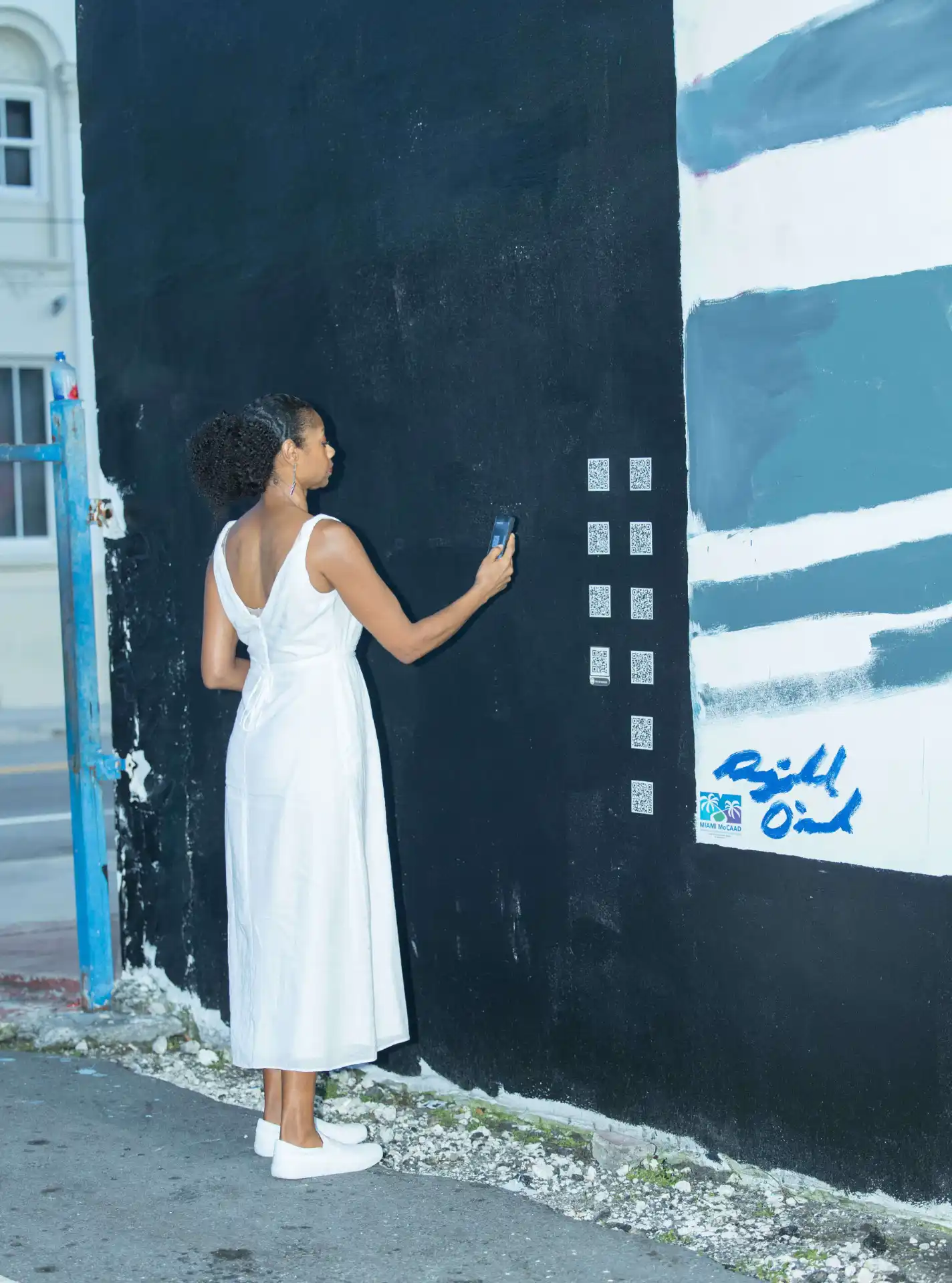 A woman in white dress using phone by a black and striped wall.