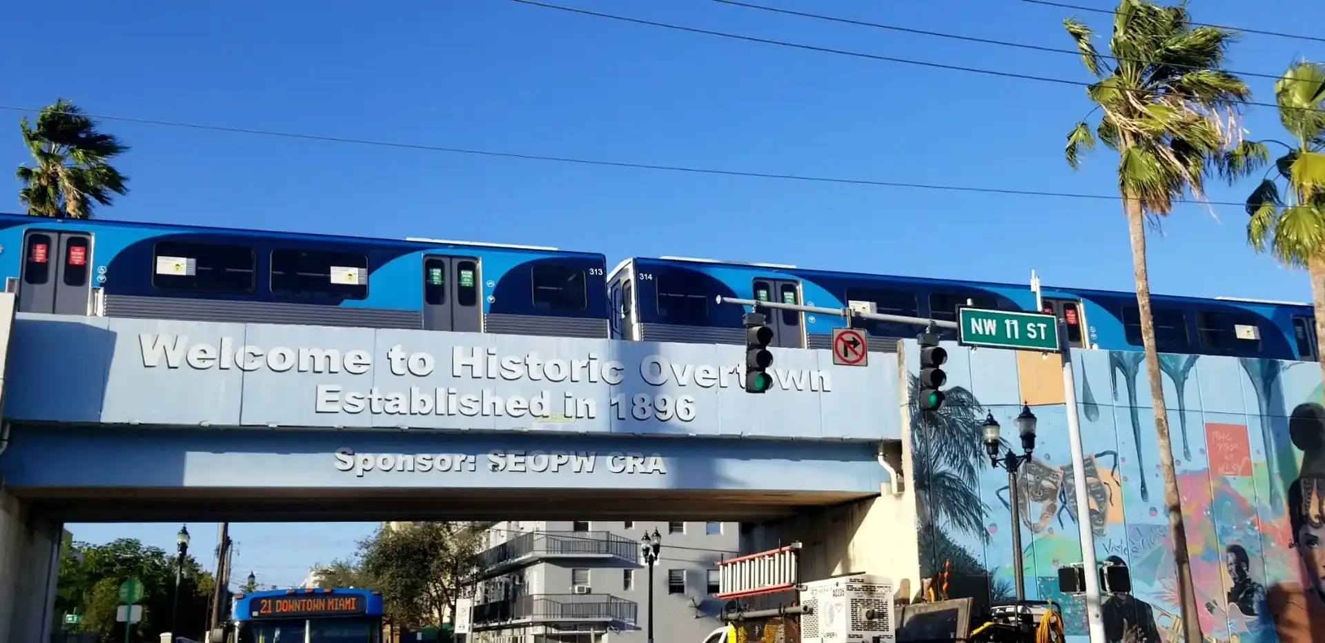 Light rail train passing over a bridge with sign reading 'Welcome to Historic Overtown Established in 1896' and street sign NW 11 St.
