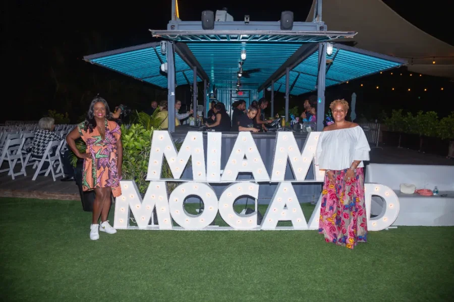 Two women posing beside large illuminated letters spelling 'MIAMI MOCA AD' at an outdoor night event with people and chairs in the background.