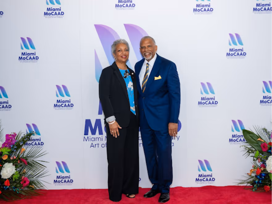Man in a blue suit and woman in black pantsuit posing on a red carpet in front of a Miami MoCAAD backdrop with floral arrangements on both sides.