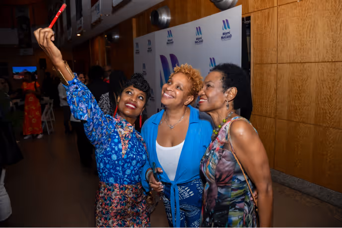 Three women smiling and posing together for a selfie at an event with Miami MoCAAD banners in the background.