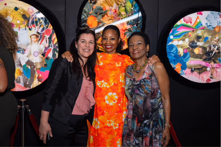 Three women smiling and posing together in front of colorful circular art pieces on a black wall.
