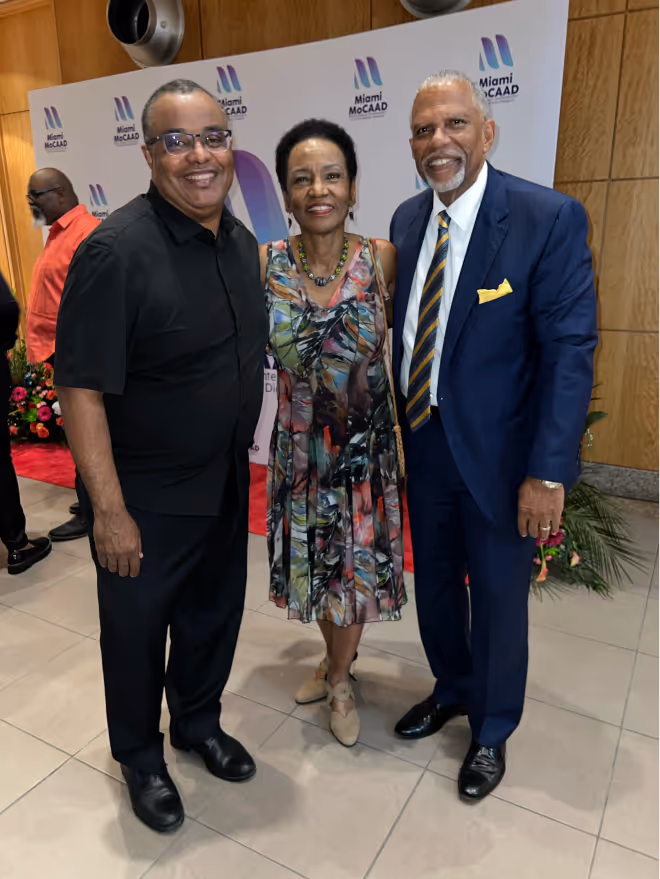 Three people posing and smiling indoors in front of a Miami MoCAAD backdrop, two men and one woman wearing formal and semi-formal attire.