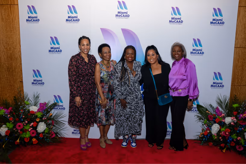 Five women standing on a red carpet in front of a Miami MoCAAD branded backdrop, flanked by colorful flower arrangements.