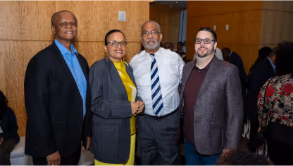 Four adults standing together smiling, three men and one woman dressed in formal and business casual attire against a wooden wall background.