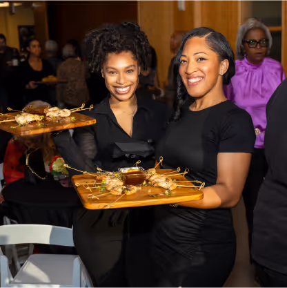 Two women smiling and holding wooden trays with skewered appetizers and dipping sauce at an indoor event.