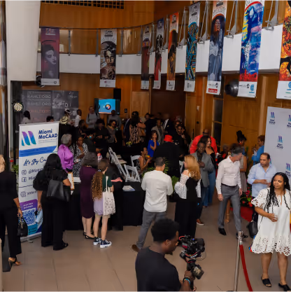 People socializing at an indoor event with Miami McCADD banners and colorful artwork hanging on the walls.