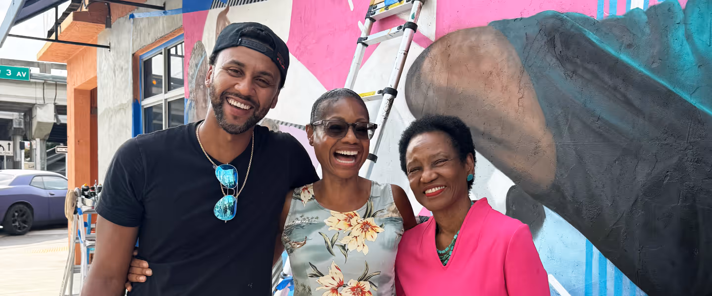 Three people smiling and standing with arms around each other in front of a colorful mural and a ladder.