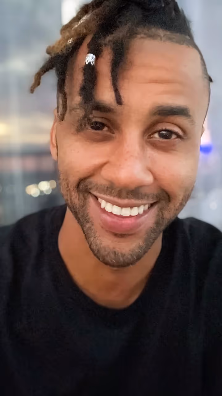 Close-up of a smiling man with dreadlocks wearing a black shirt against a blurred background.