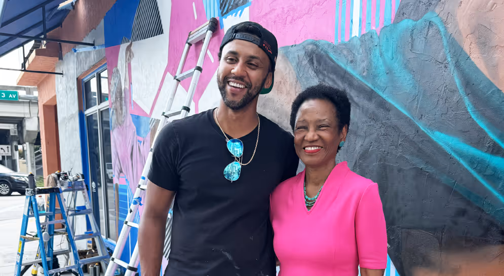 A smiling man wearing a black cap and shirt stands next to a smiling woman in a bright pink top in front of a colorful mural with ladders nearby.