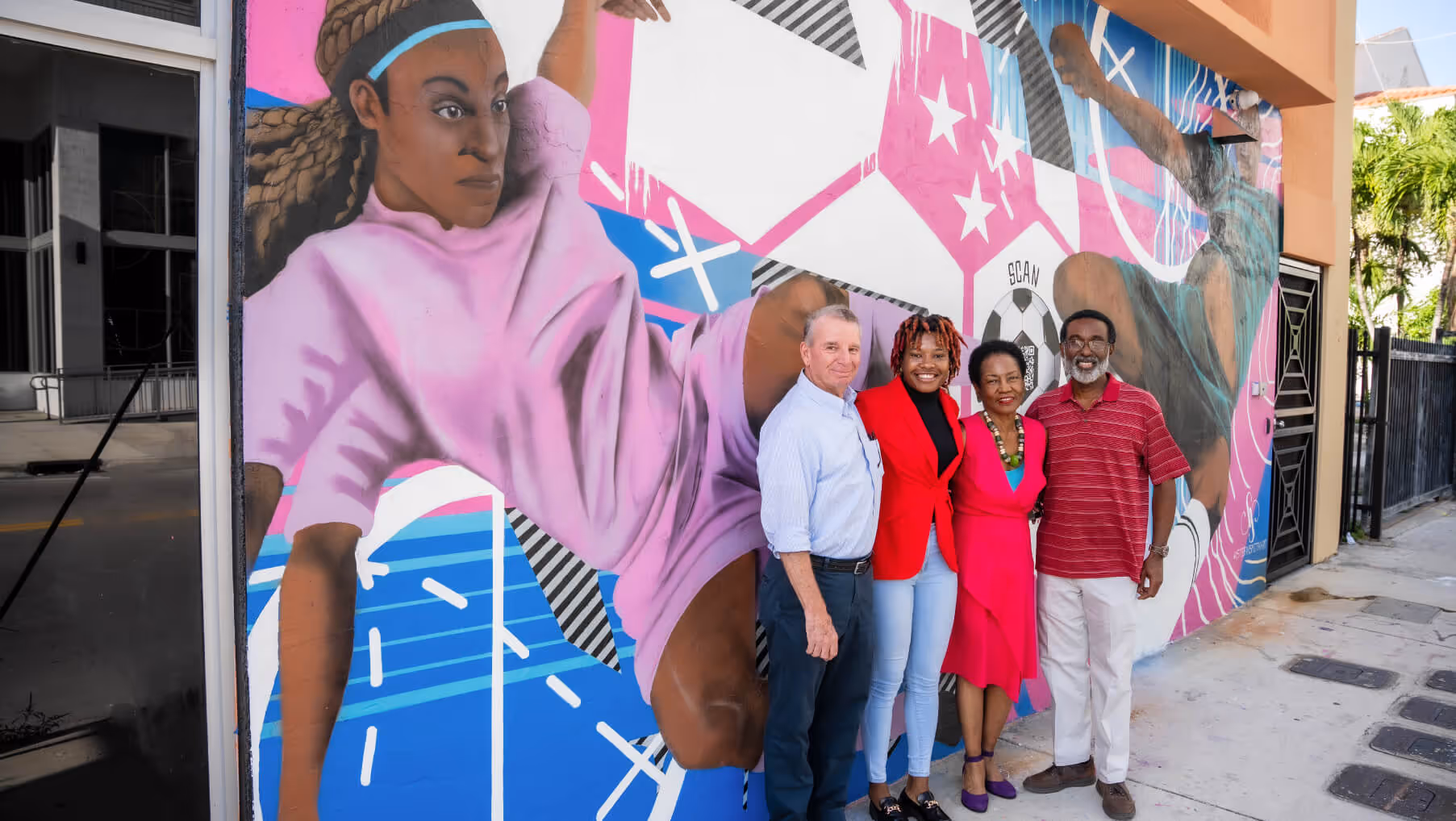 A group of four people standing and smiling in front of a colorful mural depicting two soccer players in action.
