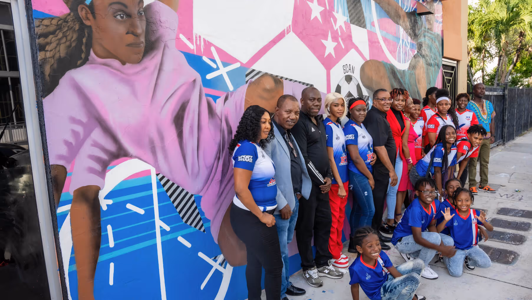Group of people, including adults and children in sports uniforms, posing in front of a colorful mural depicting a female soccer player.
