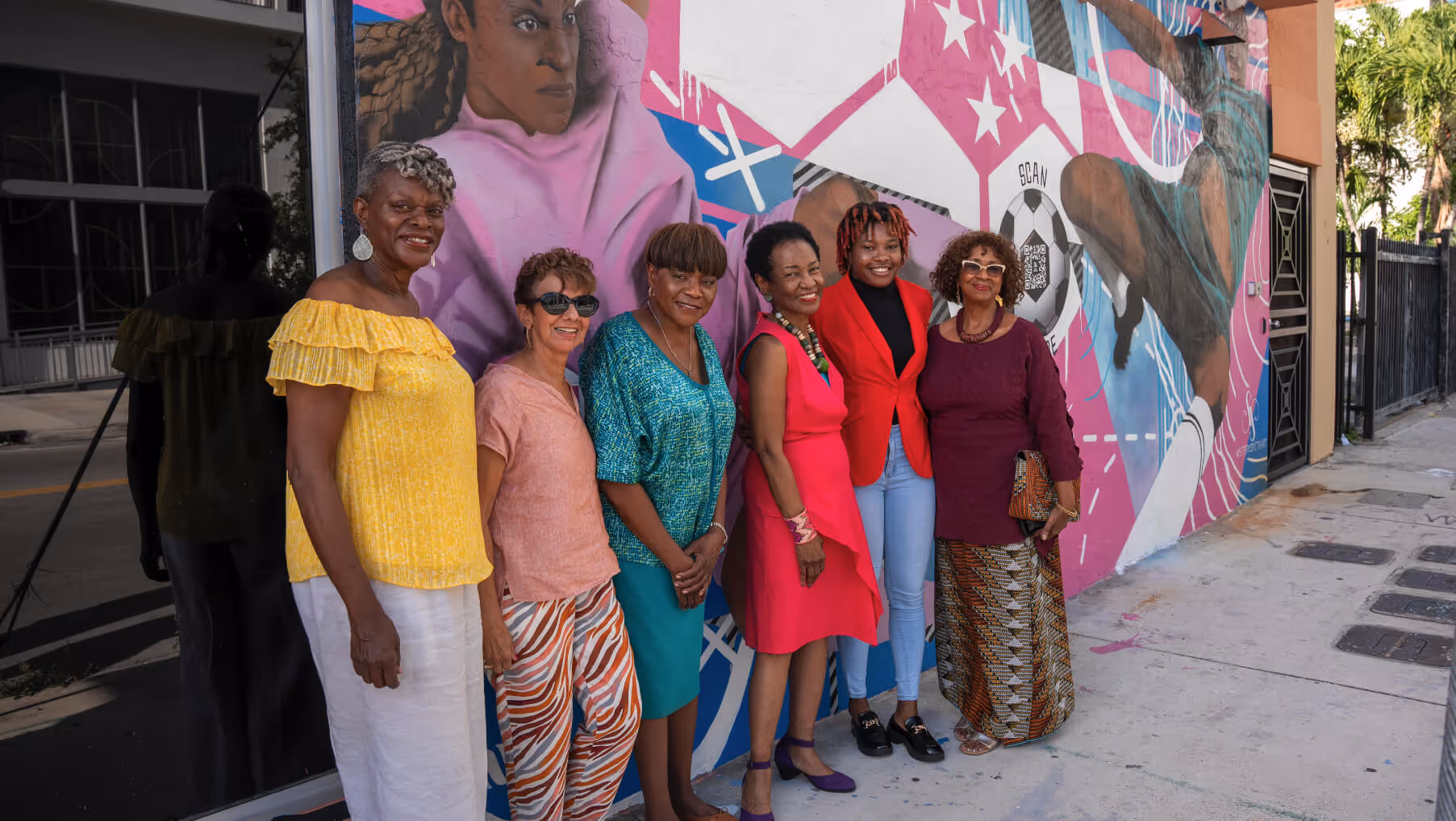 Six women standing in a row smiling in front of a colorful mural with a soccer player and geometric shapes.