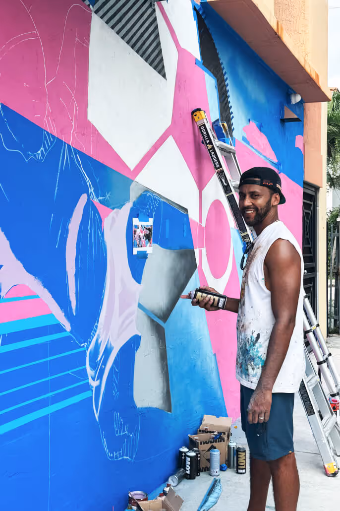 Man in a sleeveless shirt and cap painting a colorful mural with blue, pink, and white geometric shapes on a wall.