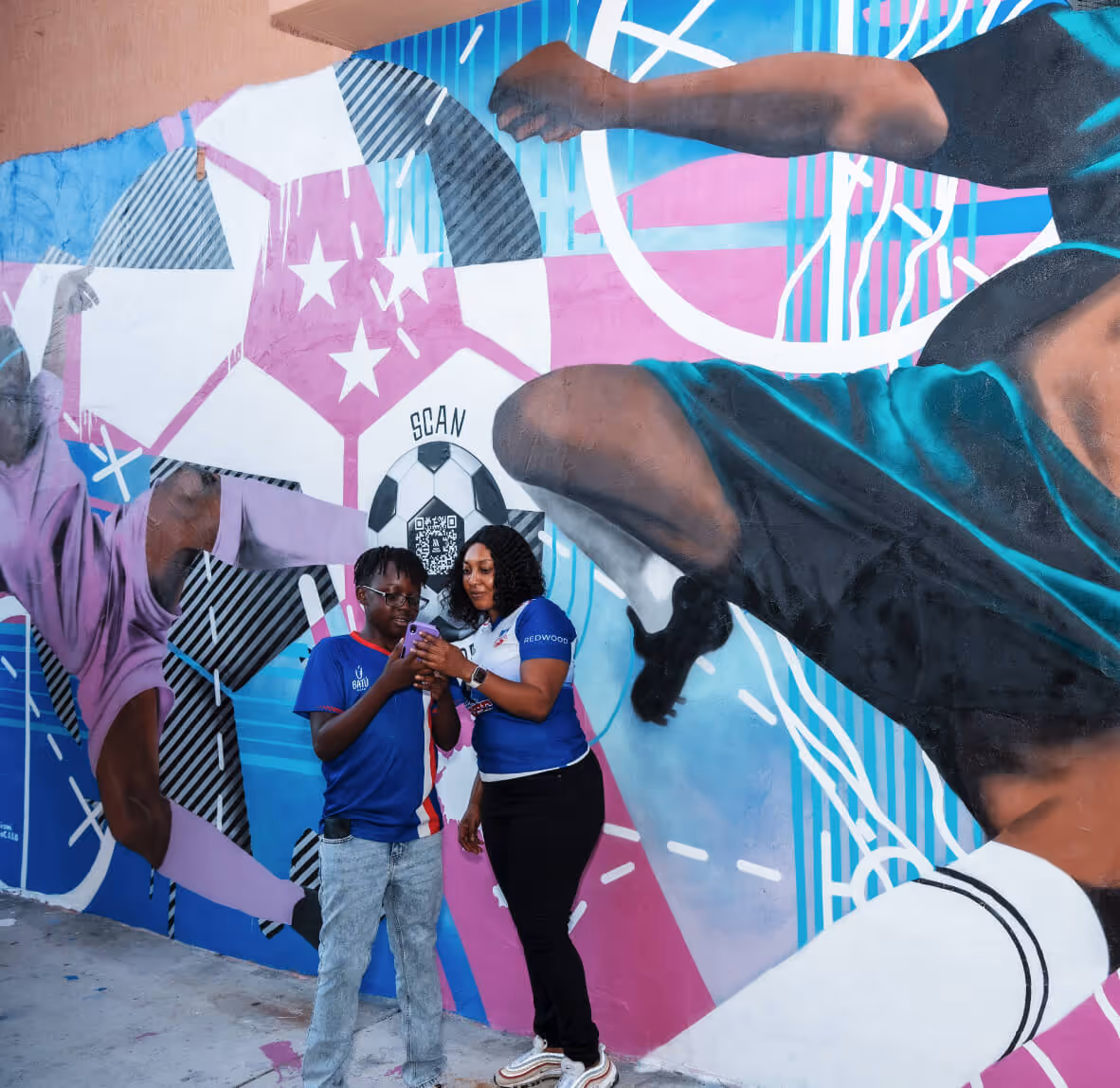 A woman and a boy standing in front of a colorful mural featuring soccer players and a soccer ball, looking at a smartphone together.