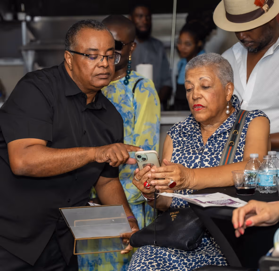 Man in black shirt pointing at a smartphone held by a woman in a blue patterned dress at a social gathering.