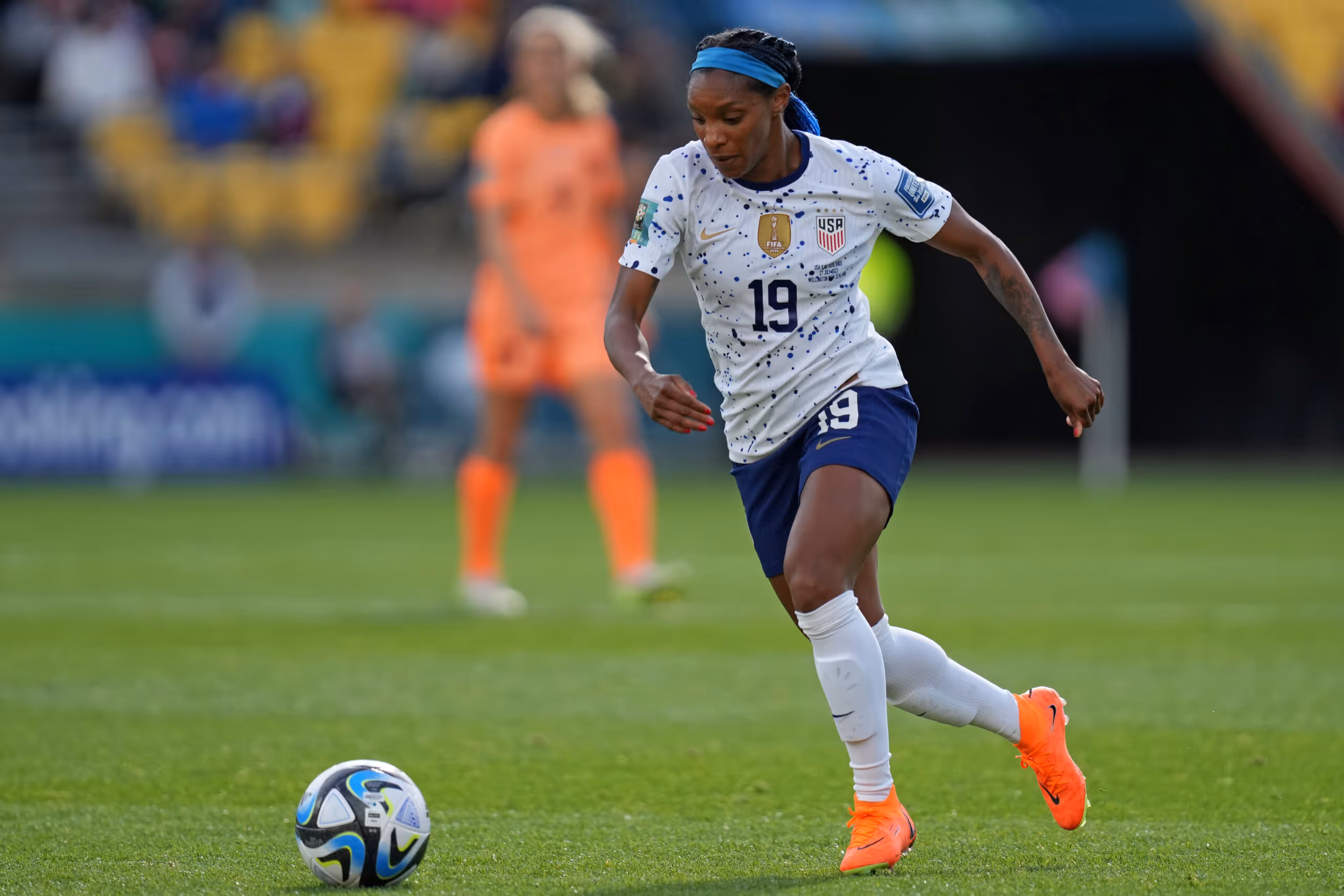 Female soccer player in white USA jersey with number 19 running towards a soccer ball on a green field.