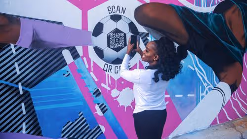 Woman scanning a QR code in the center of a painted soccer ball mural on a colorful wall.