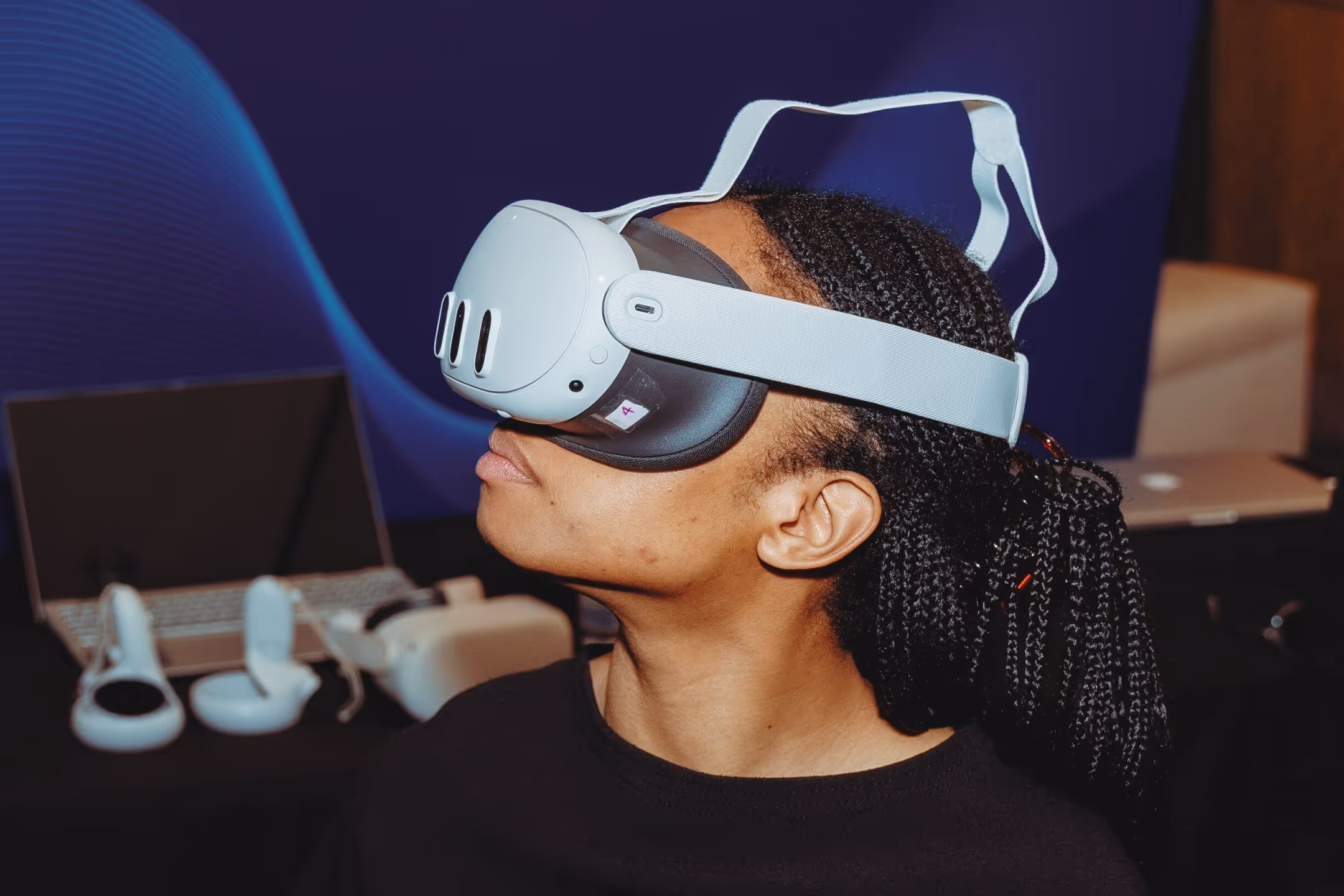 Person wearing a white virtual reality headset with braided hair pulled back, with VR controllers and a laptop in the background.