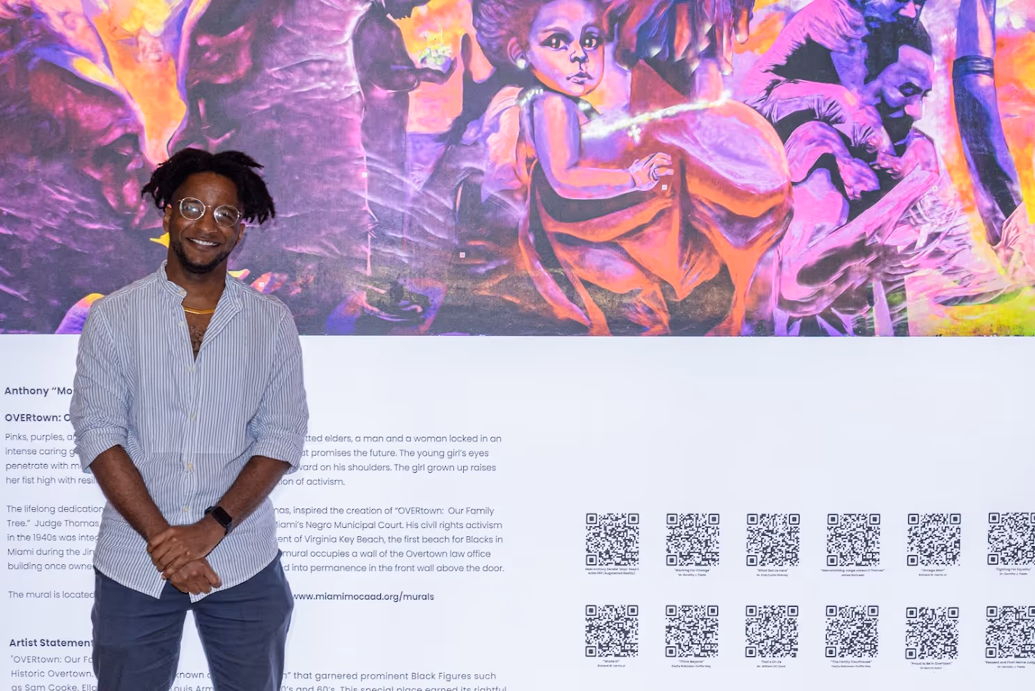 Smiling man with glasses and dreadlocks stands in front of a colorful mural featuring a child and adults in vivid purples and oranges, with text and QR codes below.