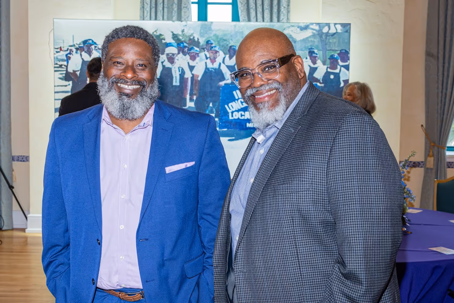 Two smiling men with gray beards and glasses, dressed in suits, standing indoors in front of a large photo of a group of people.