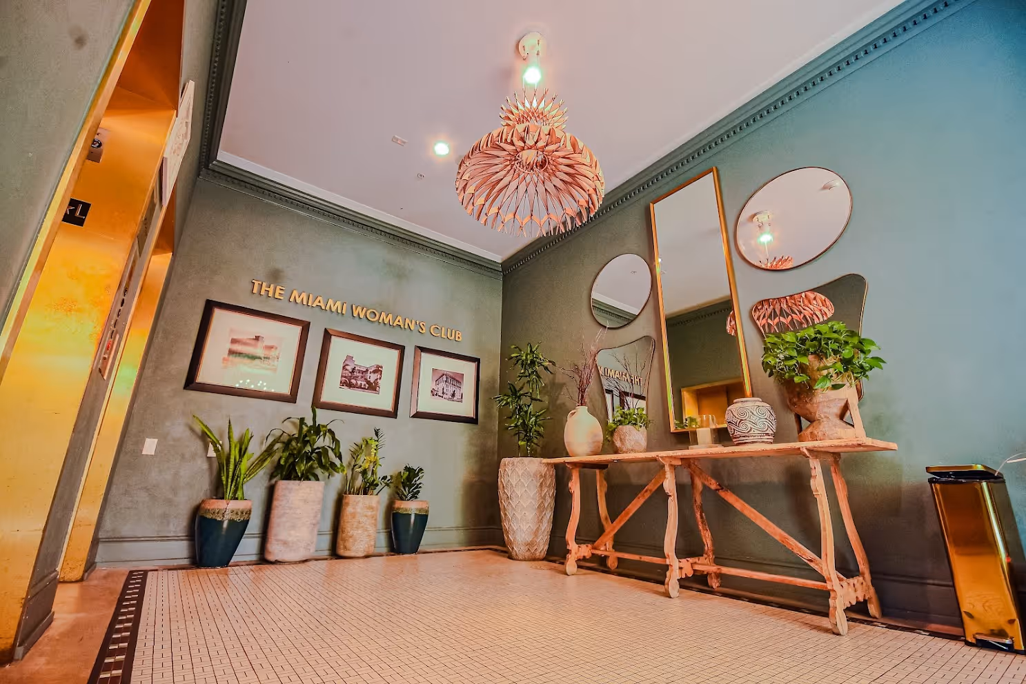 Interior of The Miami Woman's Club lobby with green walls, framed photos, a decorative wooden table with plants and vases, round and rectangular mirrors, and a hanging wooden chandelier.