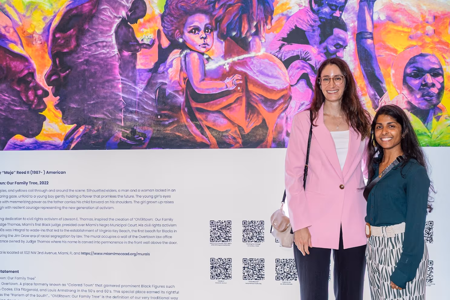 Two women smiling in front of a colorful mural featuring purple and orange hues with portraits and figures.