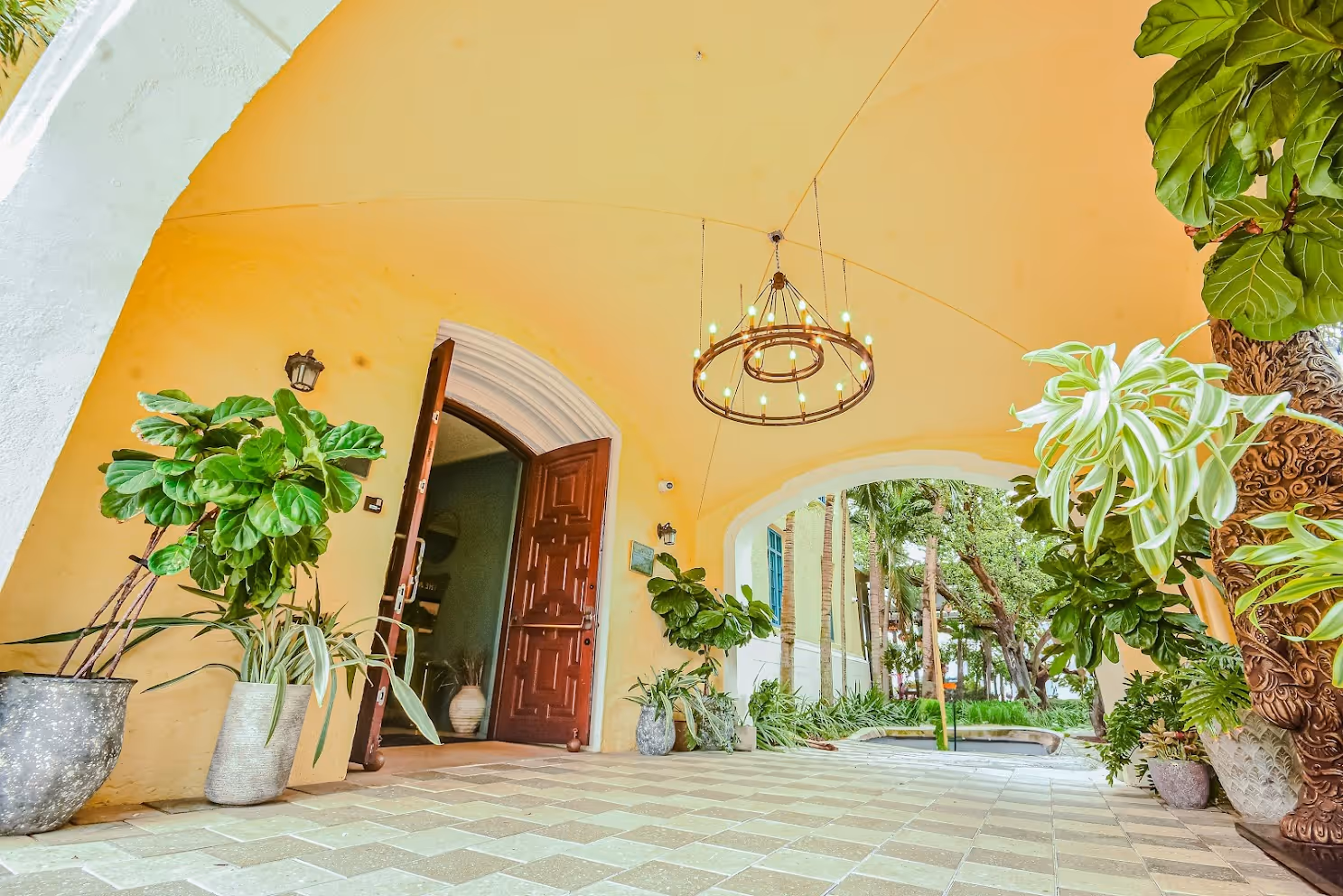Open wooden door under a yellow arched walkway with potted green plants and a circular chandelier, leading to a garden area.
