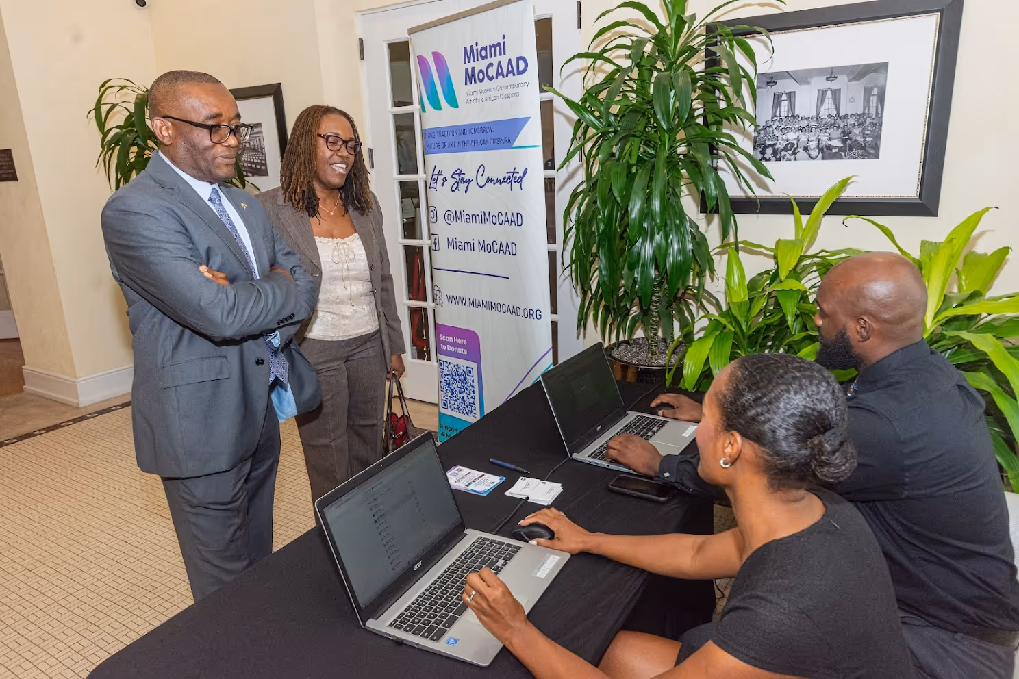 Two people standing and talking to two seated individuals working on laptops at a Miami MoCAAD event registration table indoors.
