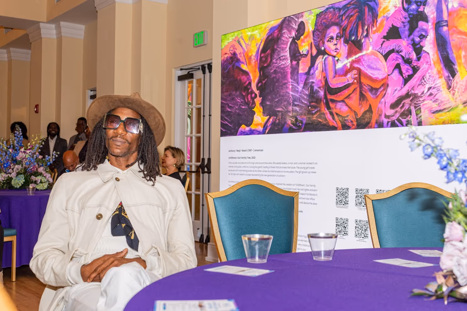 Man wearing a brown hat, large sunglasses, and white jacket sitting at a purple-decorated table in front of a colorful mural in an event space.