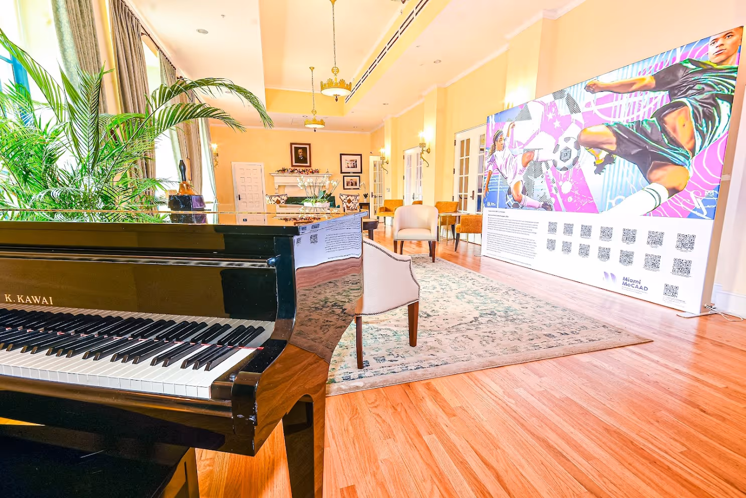 Bright room with a black Kawai piano, green plants, seating area with chairs, and a large colorful mural of soccer players on the wall.