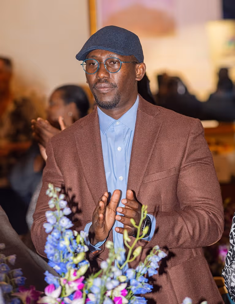 Man in round glasses, a dark cap, light blue shirt, and brown blazer clapping at an indoor event with blue and purple flowers in the foreground.