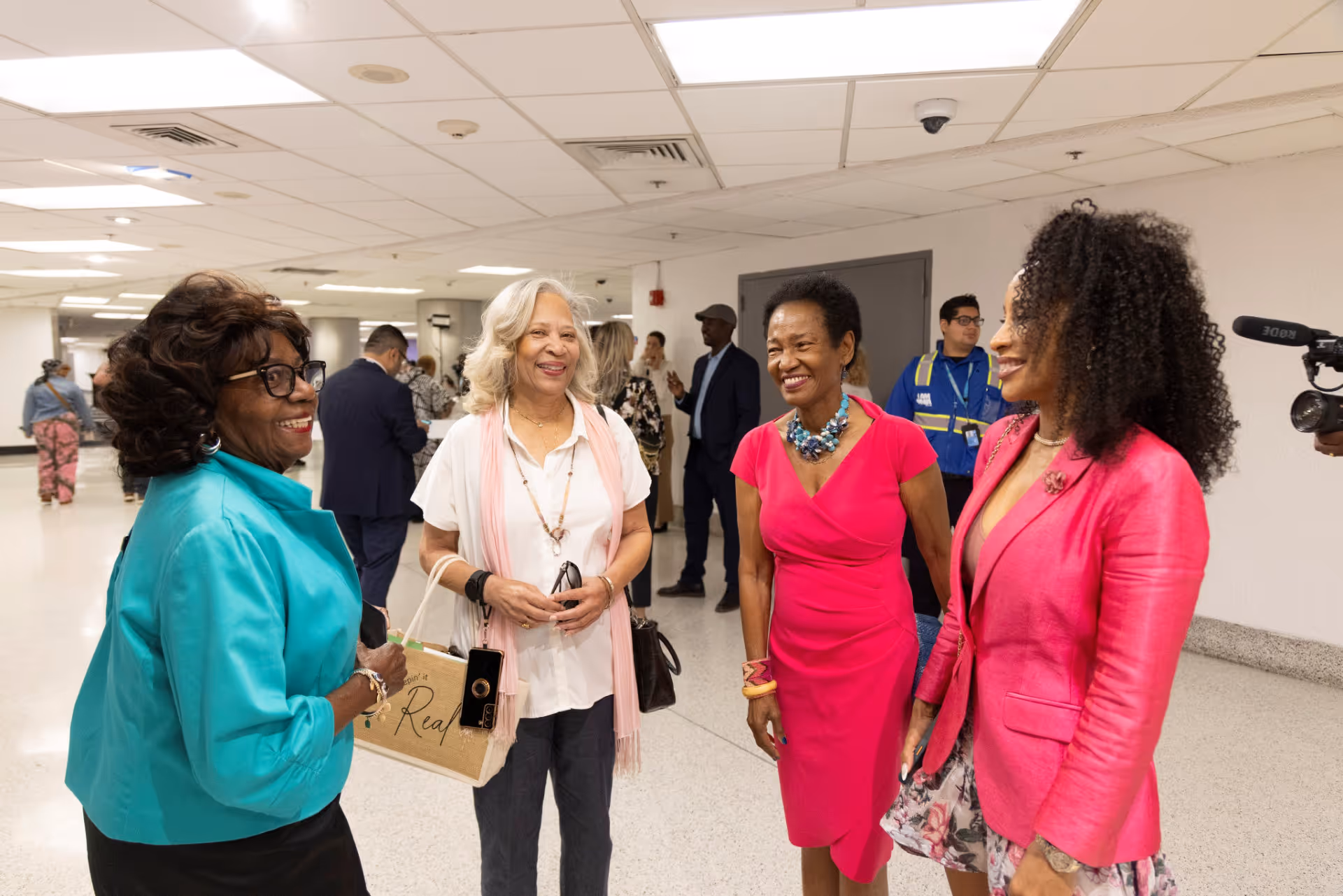 Four women smiling and chatting in a bright indoor hallway, dressed in colorful attire.