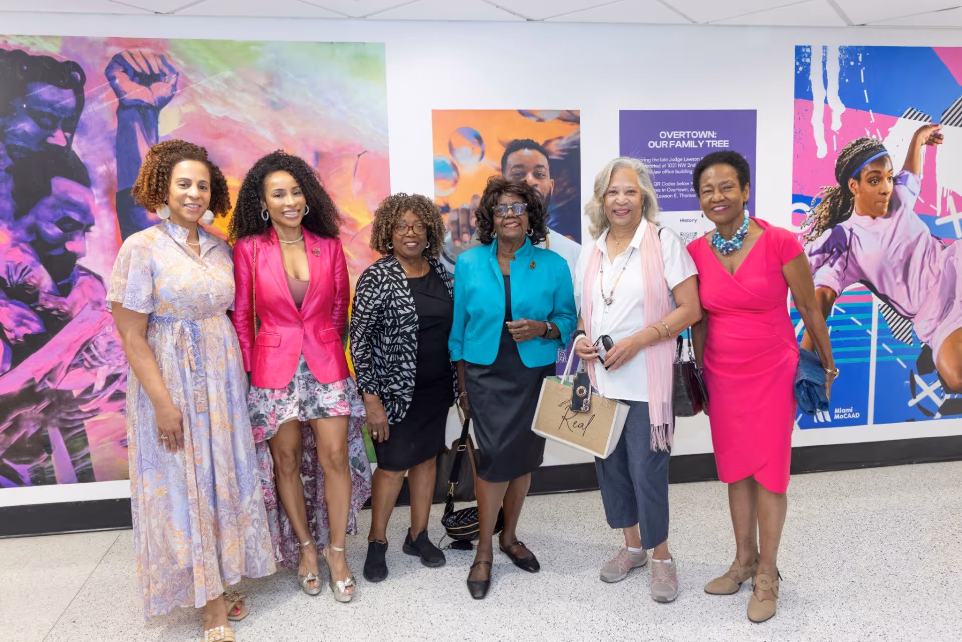 Six women standing in front of colorful murals, smiling at the camera in a gallery setting.