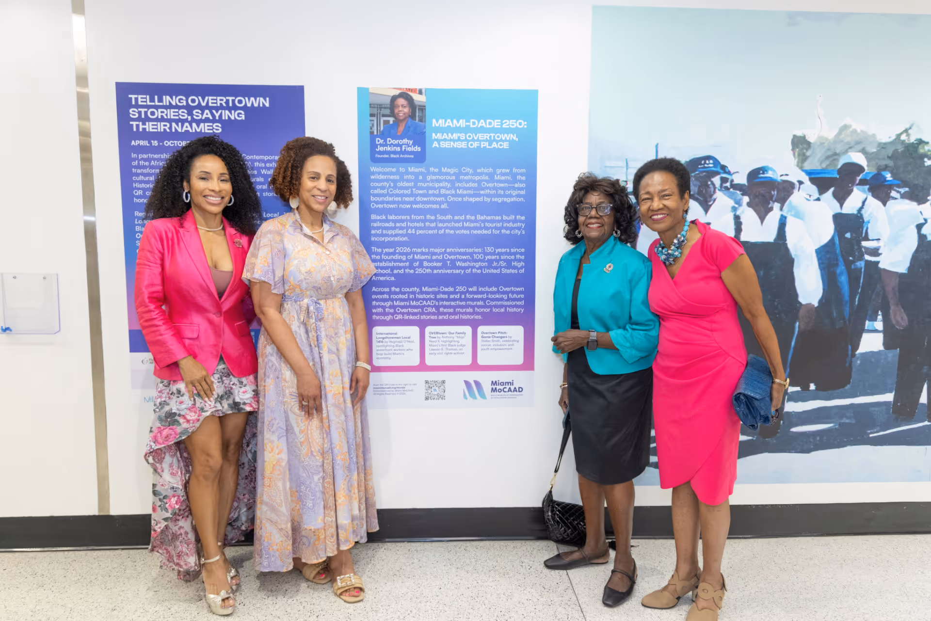 Four women standing and smiling in front of a Miami Overtown mural exhibition display.