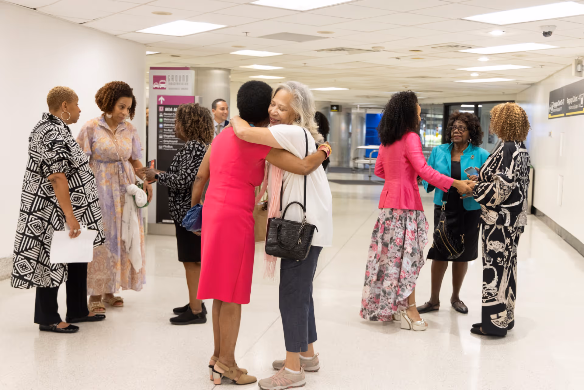 Group of women greeting and conversing in a brightly lit hallway, with two women hugging center foreground.