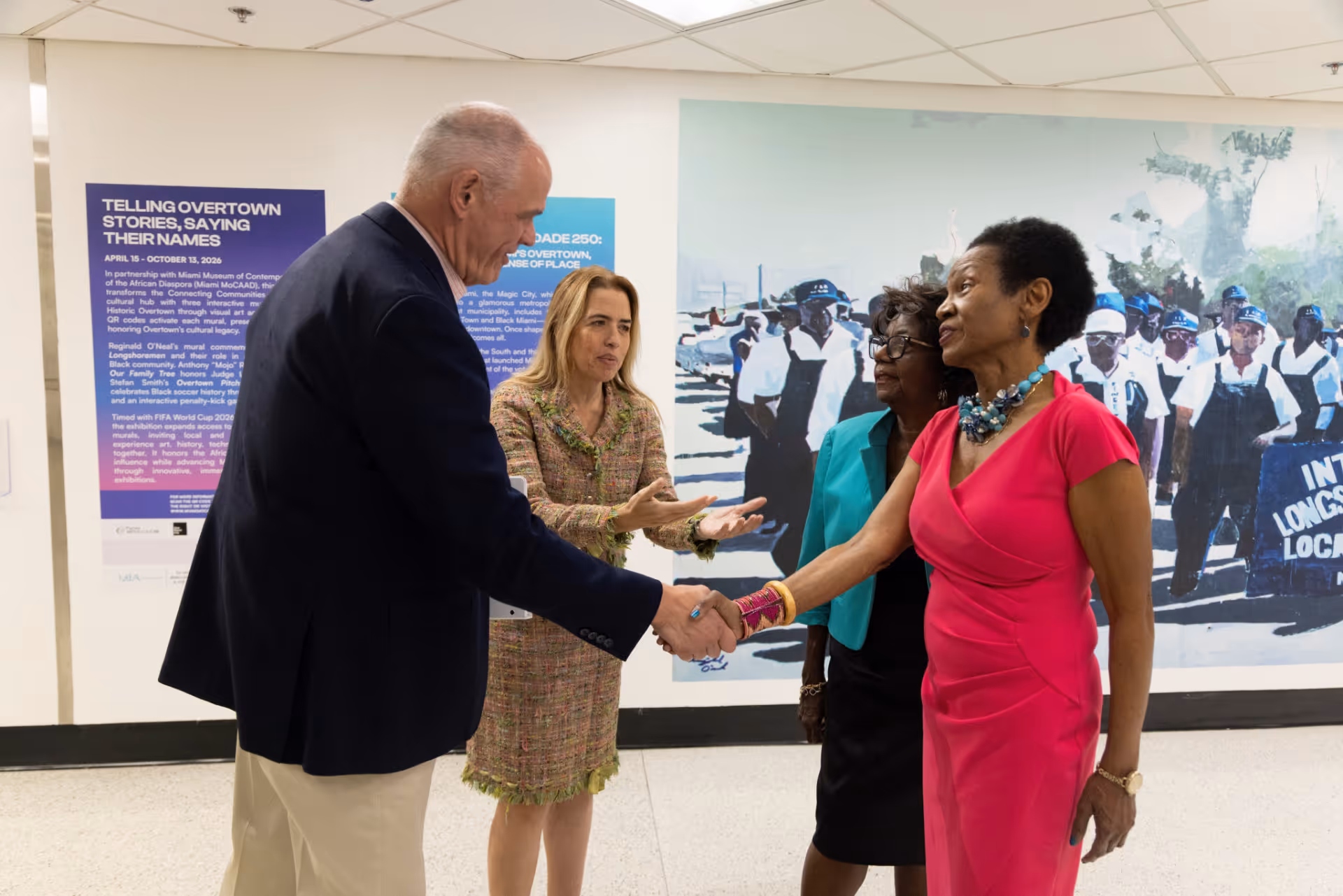 Two women and a man shake hands and talk in front of a mural at an exhibition.