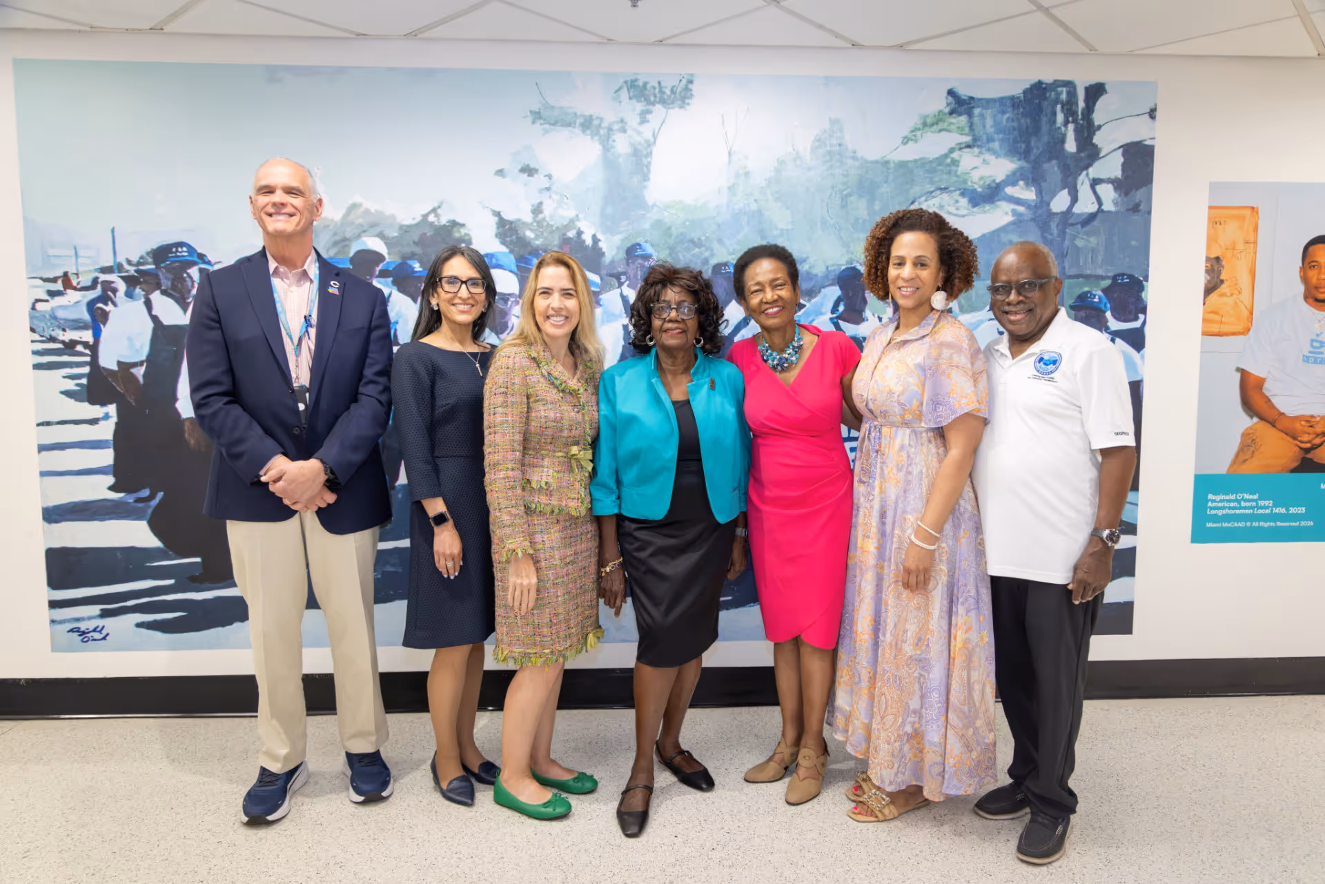 Seven people standing in front of a mural, smiling and posing for a group photo indoors.