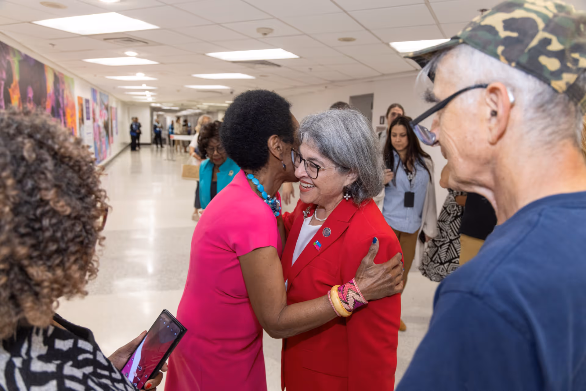 Two women warmly greeting each other with smiles and an embrace in a hallway with other people nearby.