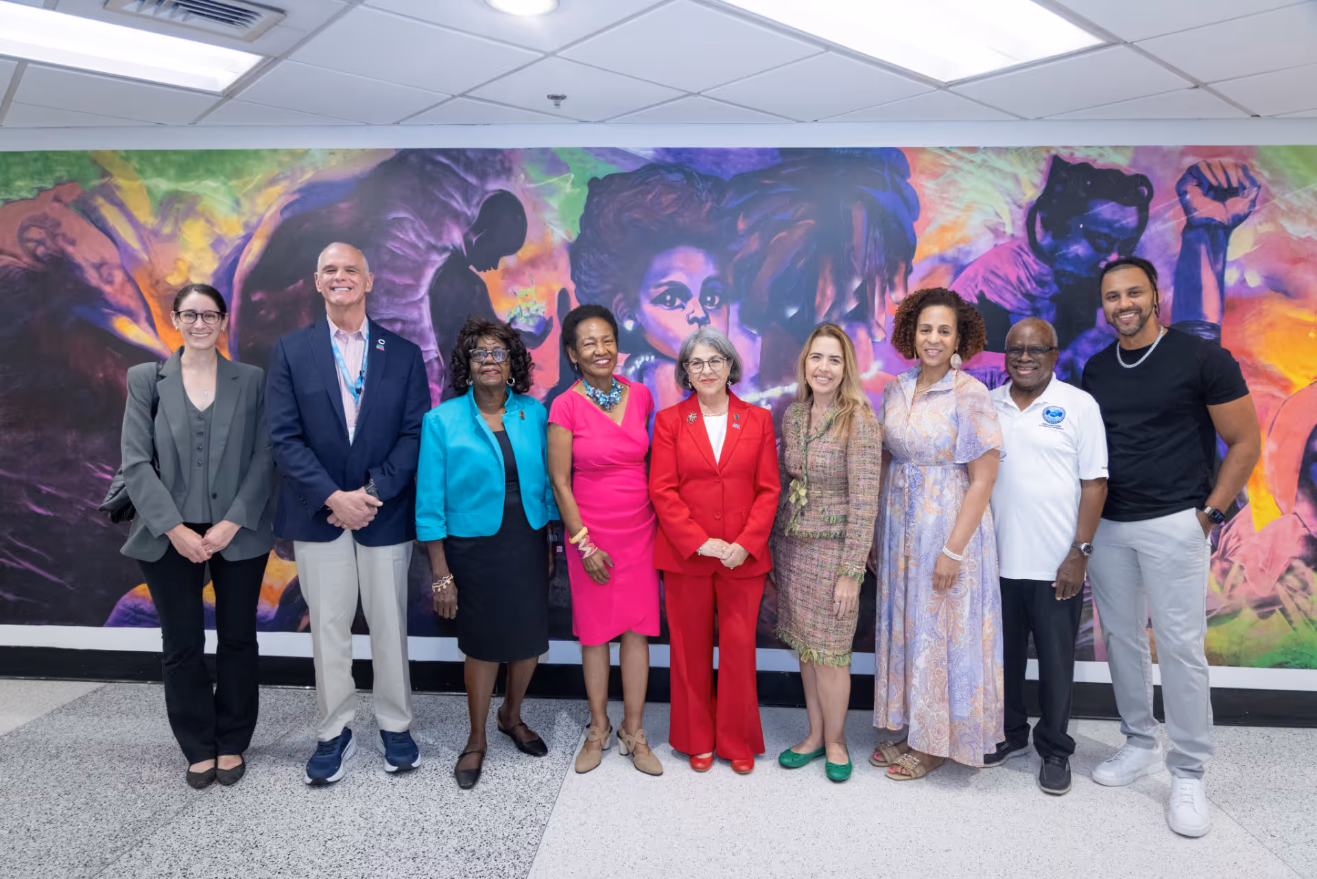 Group of nine diverse people standing indoors in front of a colorful mural with raised fist and faces.