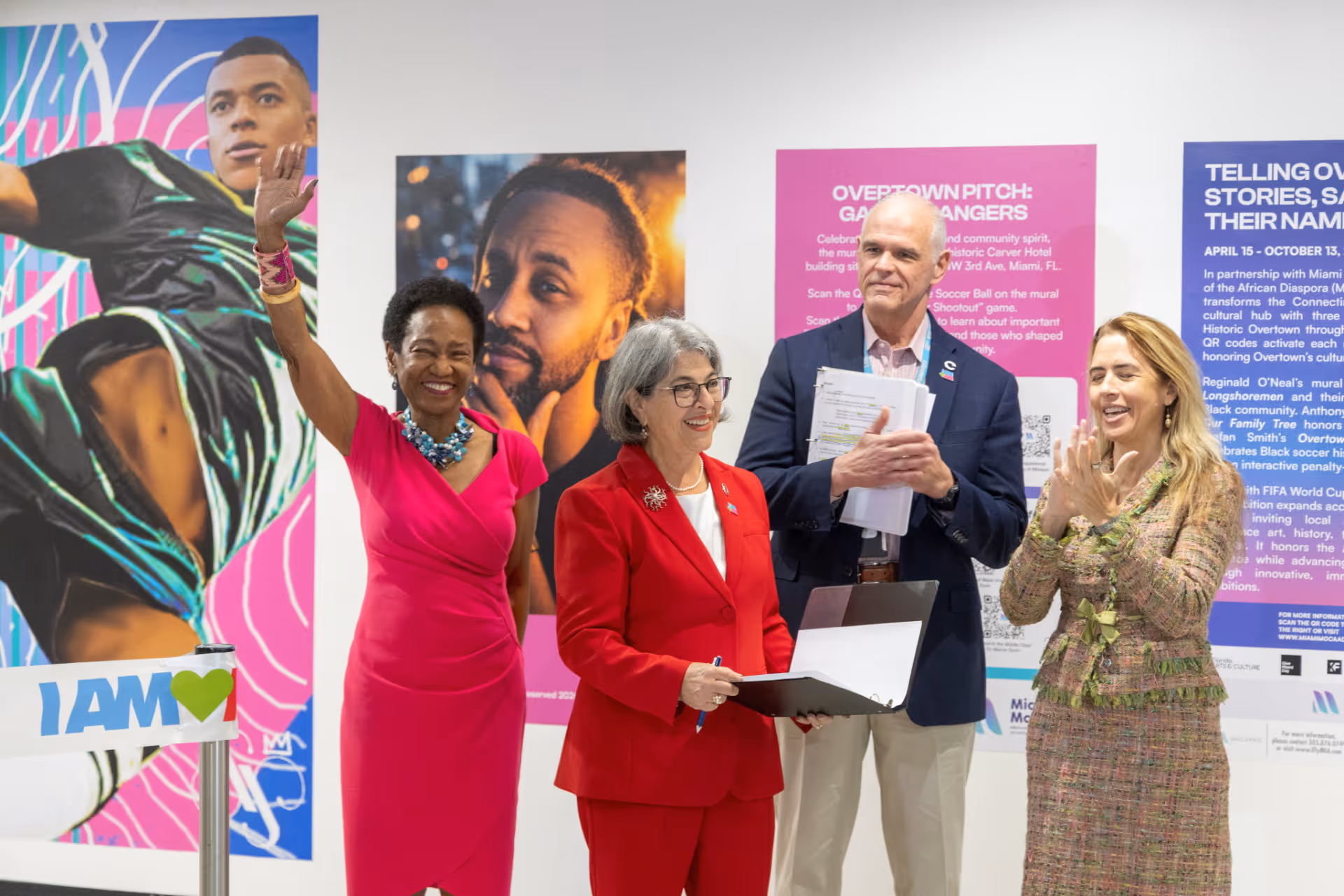 Four people standing in front of colorful posters at an event, one woman in pink waves and another claps.