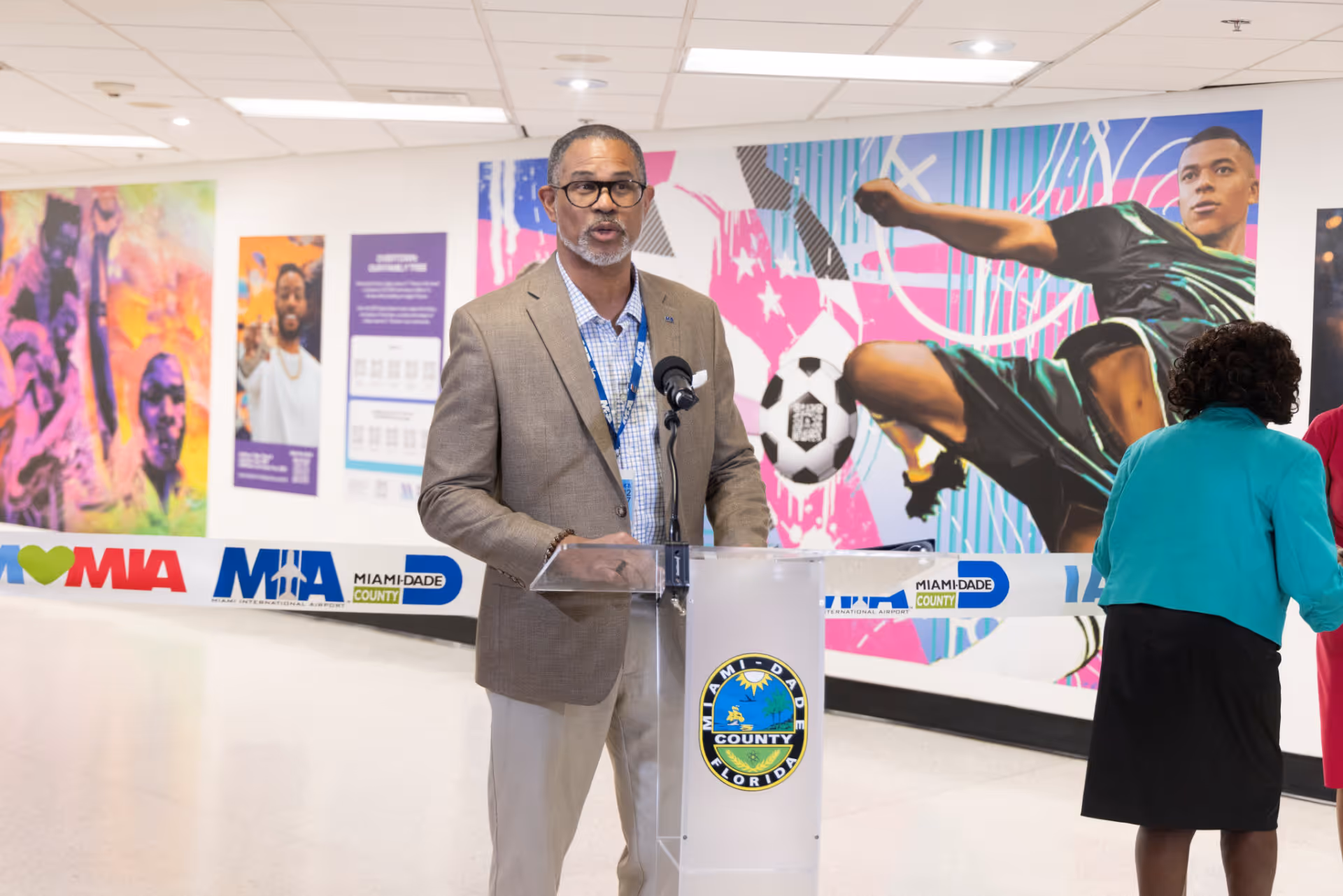 Man in glasses and brown blazer speaking at a Miami-Dade County podium with colorful mural background.
