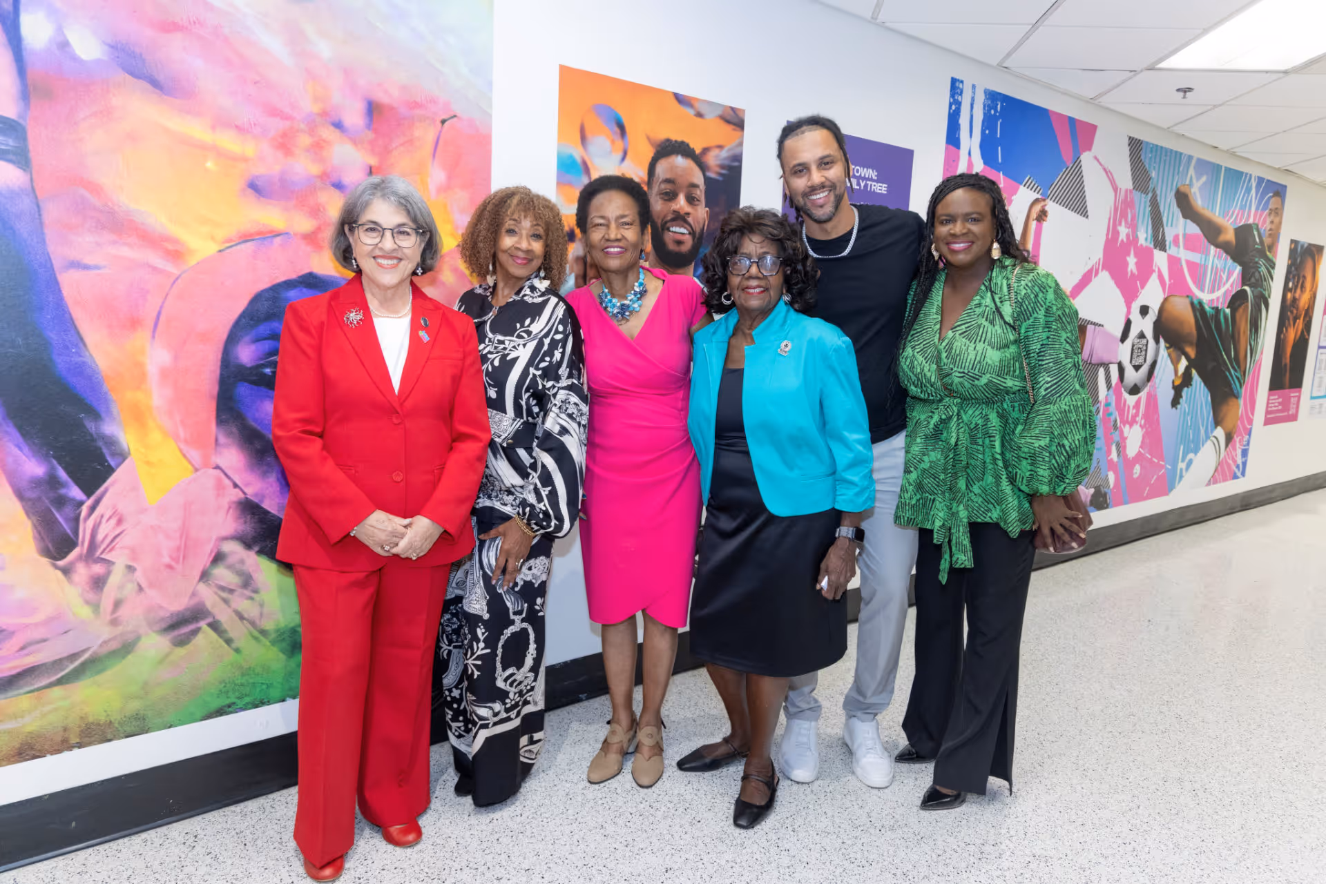 Group of six adults standing and smiling in front of colorful mural art inside a building.