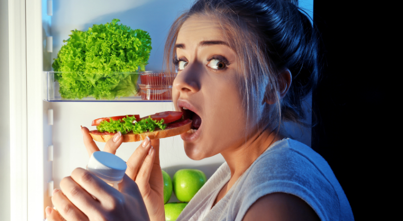 A girl eating an open sandwich in front of an open fridge 