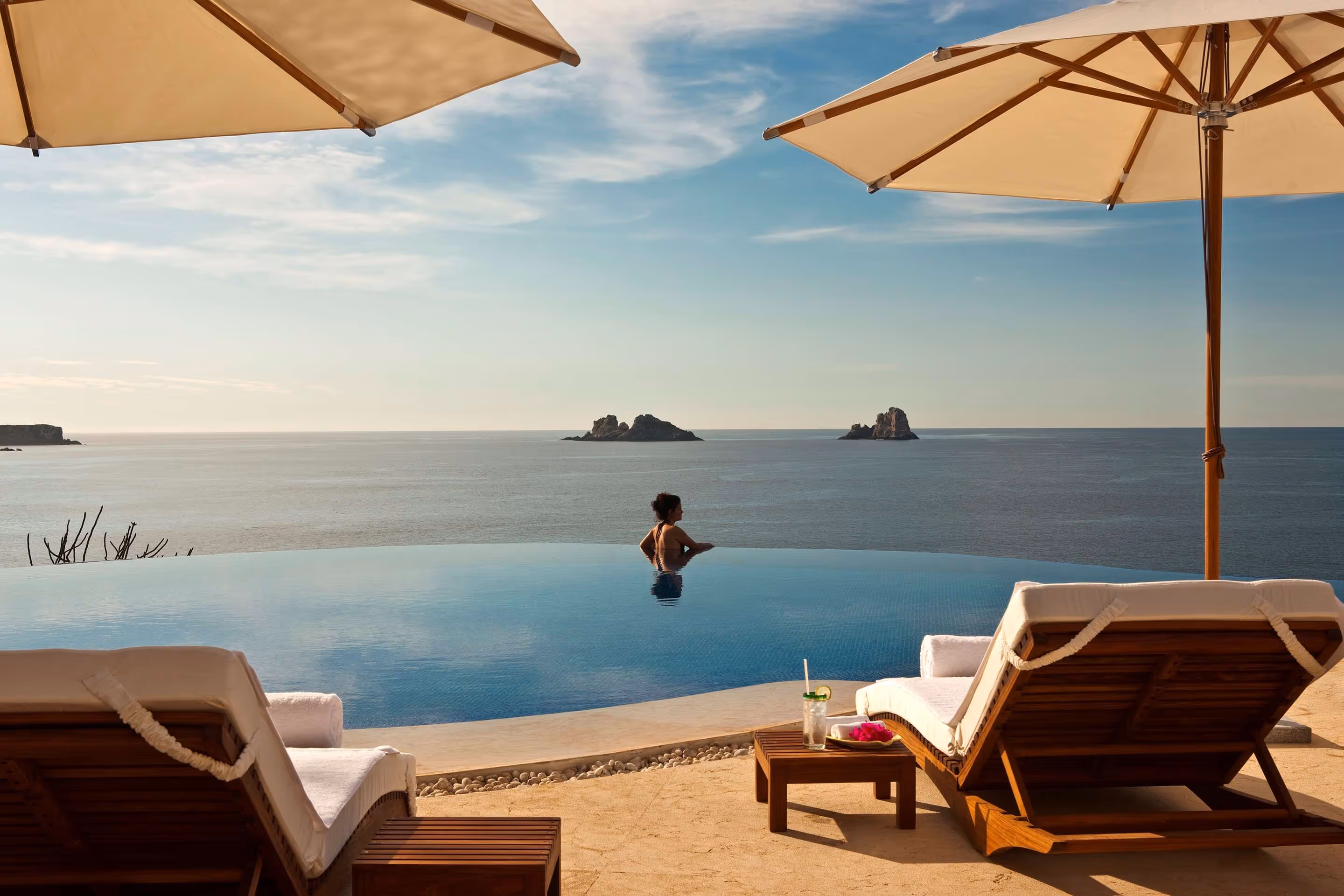 View into a hotel sun area and pool with a lady in it during a day time with a horizon of a vast blue sea and beige umbrellas.