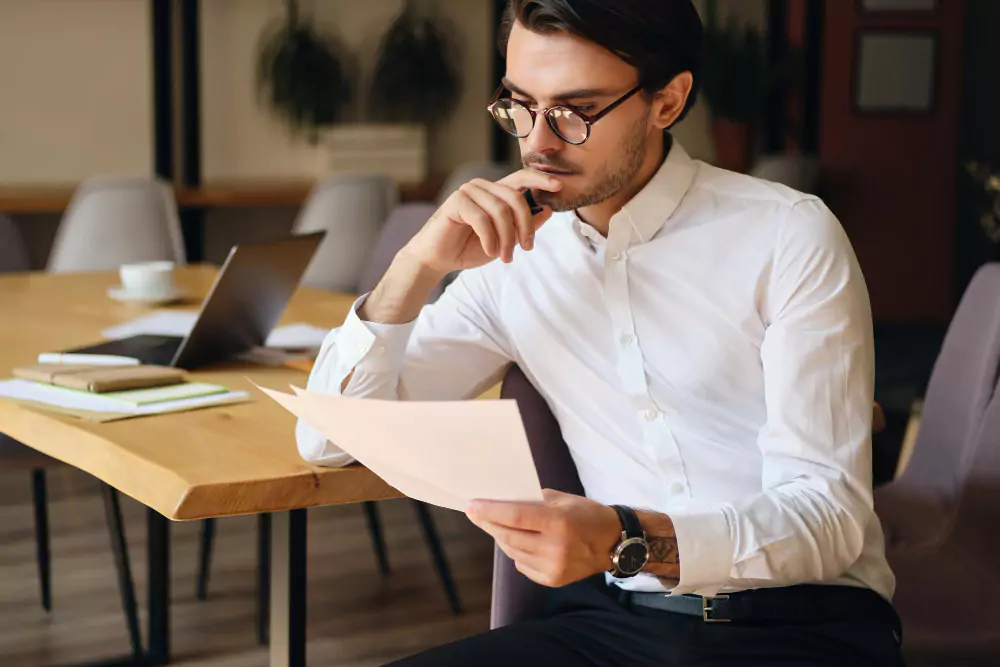 Jeune homme d'affaires sérieux portant des lunettes lisant attentivement des documents tout en travaillant dans un bureau moderne