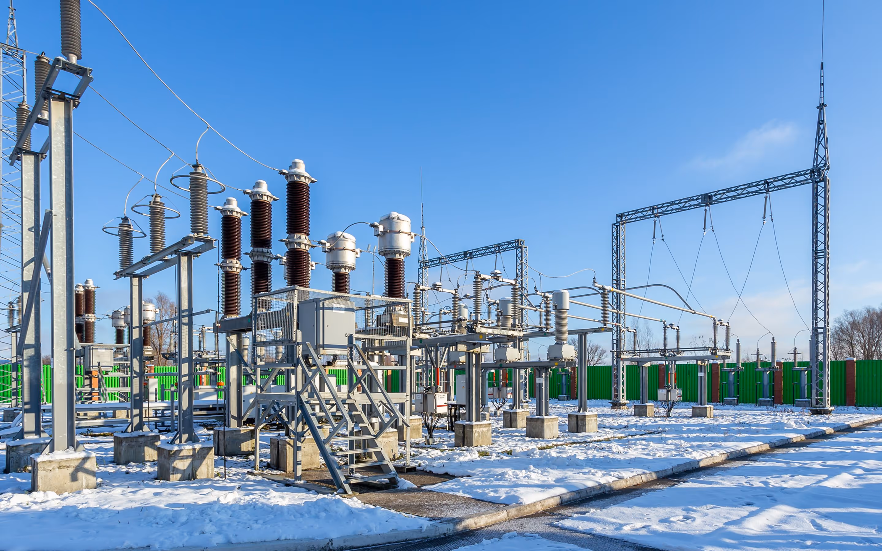 Outdoor electrical substation with transformers and high-voltage equipment on a snowy ground under a clear blue sky.
